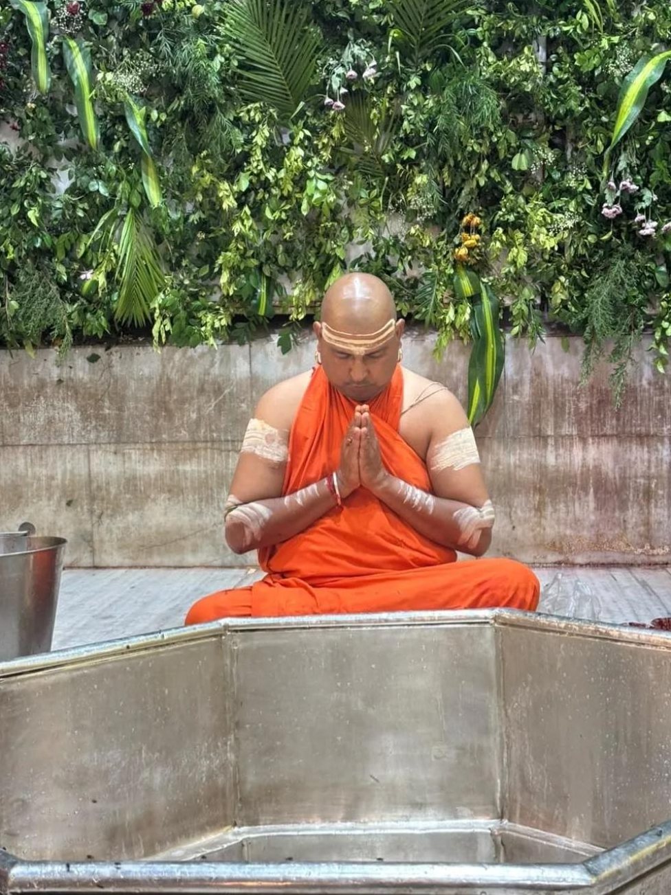 A pujari in saffron seated in prayer beside the Shivling enclosure